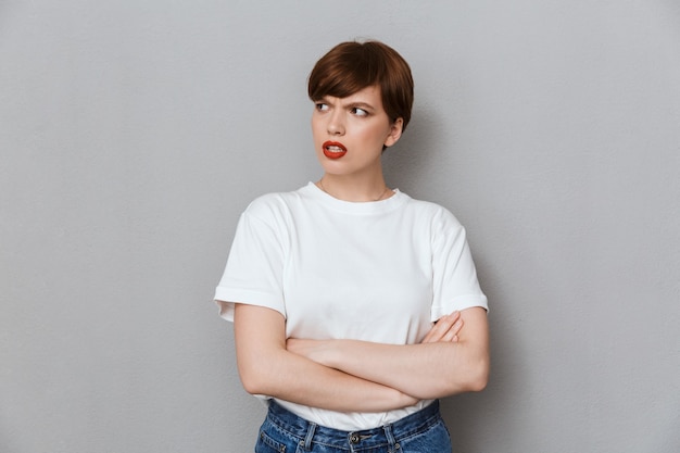 Portret van een wrokkige brunette vrouw die een casual T-shirt draagt, fronsend en staand met gekruiste armen geïsoleerd over een grijze muur