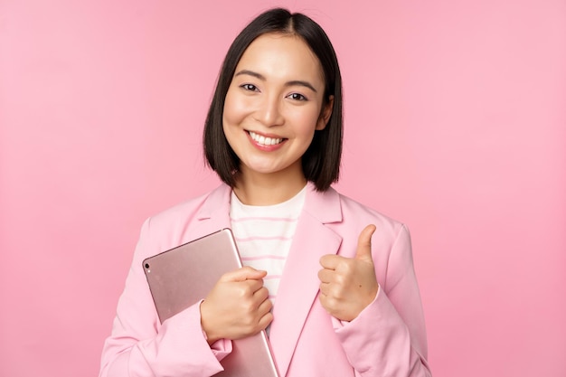 Portret van een zakelijke vrouw in een kantoor in een pak met een digitale tablet die duimen omhoog laat zien en een bedrijf aanbeveelt dat over een roze achtergrond staat