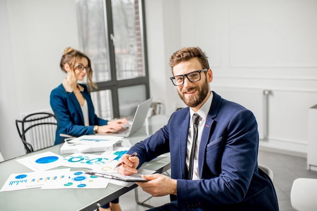 Portret van een zakenman gekleed in pak die aan documenten werkt met vrouwelijke assistent op de achtergrond in het witte kantoorinterieur