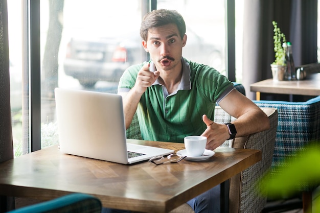 Portret van een zakenman met een groen T-shirt, die op een laptop werkt, met de vinger wijst, de richting van de camera aangeeft en er geïrriteerd uitziet, een keuze maakt. Binnen schot in de buurt van groot raam, café achtergrond.