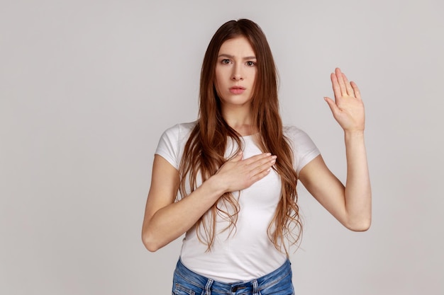 Portret van een zeer verantwoordelijke en eerlijke vrouw die belofte geeft, een plechtige gelofte aflegt in ceremoniële traditie met opgeheven hand, met een wit T-shirt. Indoor studio opname geïsoleerd op een grijze achtergrond.