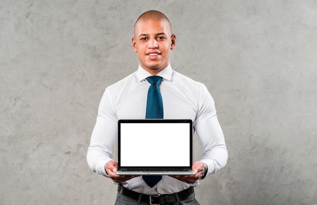 Portret van een zekere jonge zakenman die zich tegen grijze muur bevindt die laptop met het witte scherm toont