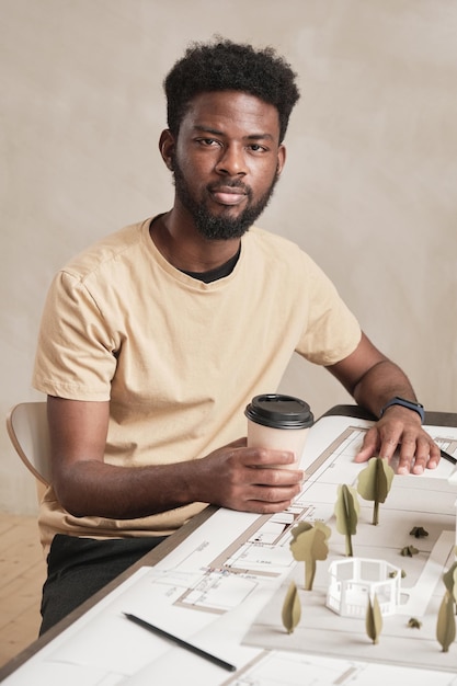 Portret van een zelfverzekerde jonge Afro-Amerikaanse architect met baard die aan tafel zit met blauwdruk en koffie drinkt op kantoor