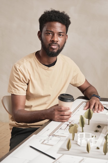 Portret van een zelfverzekerde jonge afro-amerikaanse architect met baard die aan tafel zit met blauwdruk en koffie drinkt op kantoor