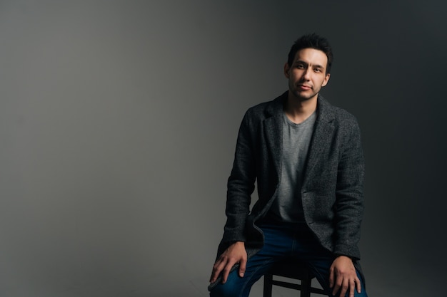 Portret van een zelfverzekerde jonge man met een stijlvolle lange jas zittend in een stoel op een zwarte geïsoleerde achtergrond die naar de camera kijkt. Studio shot van serieuze blanke man poseren tegen donkere muur.