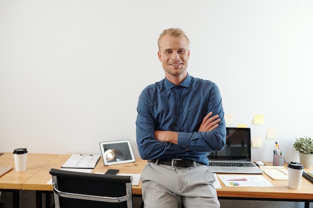 Portret van een zelfverzekerde jonge ondernemer die op een bureau leunt, armen kruist en naar de camera glimlacht