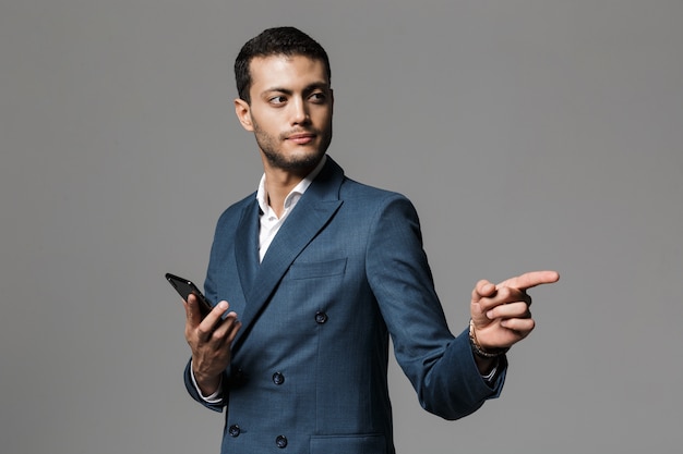 Portret van een zelfverzekerde jonge zakenman gekleed in pak staande geïsoleerd over grijze muur, met mobiele telefoon, wegwijzend