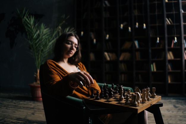 Portret van een zelfverzekerde jonge zakenvrouw met een elegante bril die schaak zet terwijl ze in een leunstoel in een donkere bibliotheekkamer zit