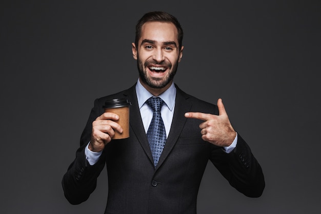 Portret van een zelfverzekerde knappe zakenman die een kostuum draagt dat zich geïsoleerd bevindt, afhaalmaaltijdenkoffie houdt