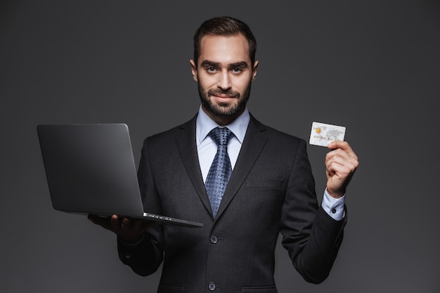 Portret van een zelfverzekerde knappe zakenman die geïsoleerde kostuum draagt, laptop computer houdt, die creditcard toont