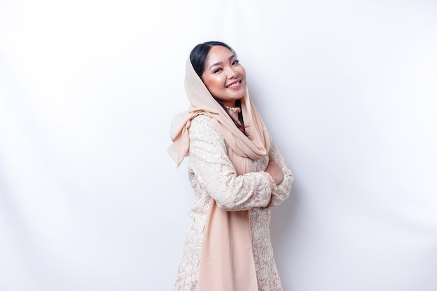 Portret van een zelfverzekerde lachende Aziatische moslimvrouw die hijab draagt en met de armen over elkaar staat en naar de camera kijkt geïsoleerd op een witte achtergrond