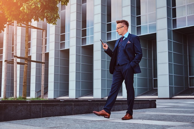 Portret van een zelfverzekerde stijlvolle zakenman gekleed in een elegant pak met een smartphone terwijl hij buiten staat tegen een wolkenkrabberachtergrond.