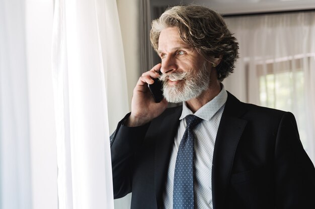 Portret van een zelfverzekerde volwassen zakenman in een zwart pak die op een smartphone praat terwijl hij bij het raam in een hotelappartement staat