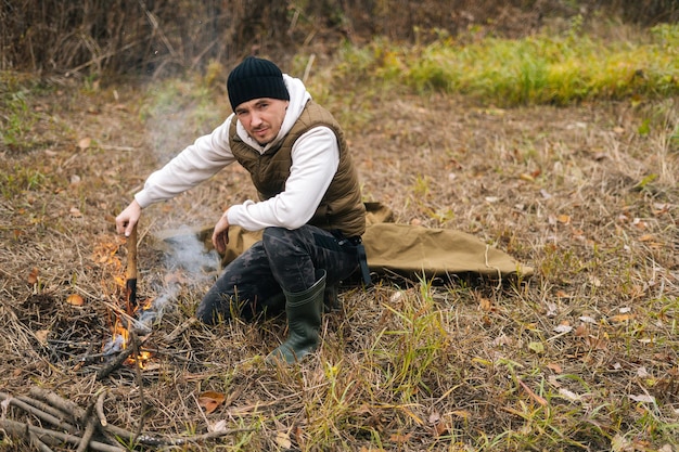 Portret van een zelfverzekerde wandelaar die warme kleren draagt en in de buurt van een brandend kampvuur zit en een kleine schop vasthoudt terwijl hij naar de camera kijkt