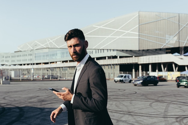 Portret van een zelfverzekerde zakenman in een klassiek pak met een videogesprek met een zakenpartner die een mobiele telefoon gebruikt die in de buurt van een modern kantoorgebouw en een witte auto staat. Technologieën concept.