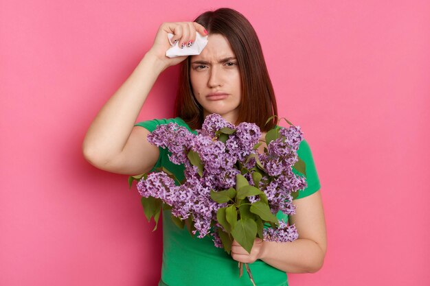 Portret van een ziek, verdrietig jong meisje met bruin haar, gekleed in een groen casual t-shirt met boeket van lila bloemen met hoofdpijn, loopneusallergie, poseren geïsoleerd over roze muur achtergrond