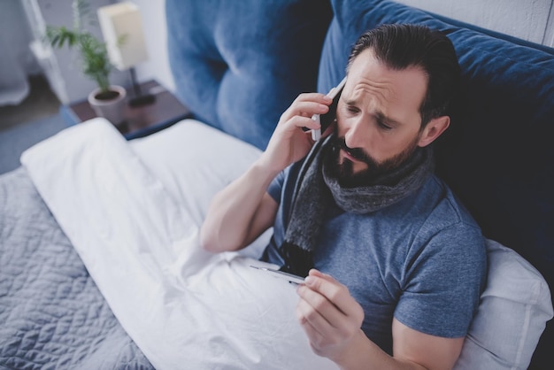 Portret van een zieke man die een thermometer vasthoudt en een dokter belt terwijl hij op het bed zit