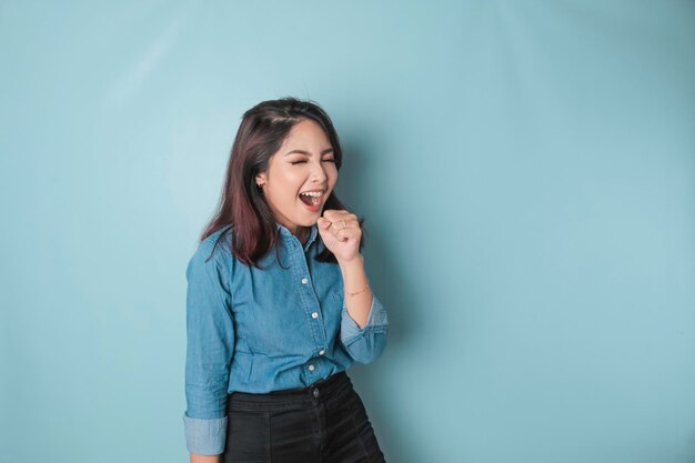 Portret van een zorgeloze Aziatische vrouw die plezier karaoke zingt in de microfoon terwijl ze over een blauwe achtergrond staat