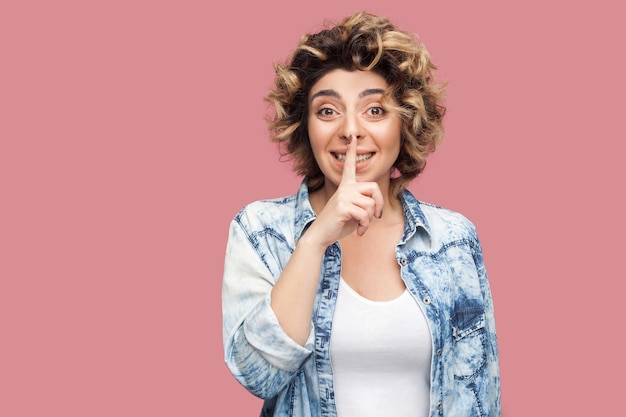 Portret van een zorgwekkende jonge vrouw met krullend kapsel in een casual blauw shirt dat staat, geheim vertelt en naar de camera kijkt met de vinger op het geheime teken van de lippen. indoor studio opname, geïsoleerd op roze achtergrond.