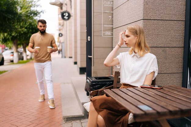 Portret van een zorgzame jonge man die koffie brengt naar een charmante blonde vriendin die op zomerdag aan tafel zit in een straatcafé