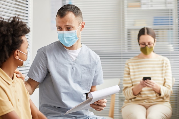 Portret van een zorgzame mannelijke arts die praat met een jongen die in de rij staat te wachten in de medische kliniek, beide met maskers, kopieer ruimte