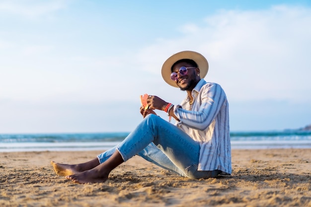 Portret van een zwarte etnische man geniet van zomervakantie op het strand en eet een watermeloen Zittend op het zand