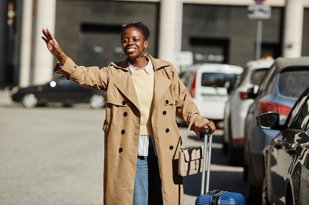 Portret van een zwarte jonge vrouw met een koffer die een taxi aanhoudt in de stadsstraat en glimlacht