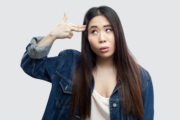 Portret van eenzame depressieve mooie brunette Aziatische jonge vrouw in casual blauw denim jasje met make-up staande met pistool pistool op hoofd. indoor studio opname, geïsoleerd op lichtgrijze achtergrond.