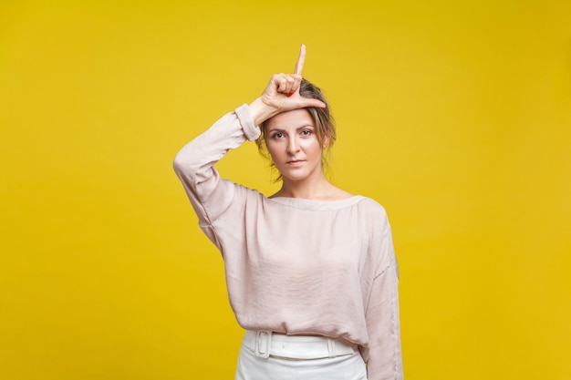 Portret van ernstig overstuur vrouw met blond haar in casual beige blouse staande met verliezer gebaar met vingers op haar hoofd kijken camera binnen studio opname geïsoleerd op gele achtergrond