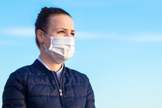 Portret van ernstig peinzend nadenkend meisje, jonge mooie vrouw in medisch steriel beschermend masker op haar gezicht op blauwe hemelachtergrond, exemplaarruimte. coronavirus, covid-19, epidemie, virusconcept.