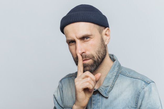 Portret van ernstige bebaarde man met aantrekkelijke look, houdt vinger op lippen, kijkt met geheime uitdrukking