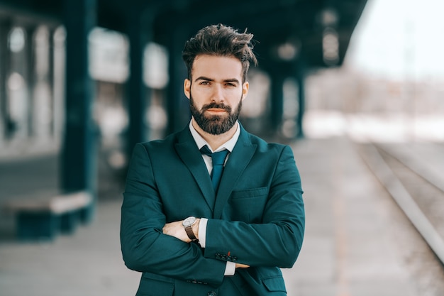 Portret van ernstige Kaukasische bebaarde zakenman in formele slijtage staande op treinstation met gekruiste armen.