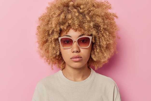 Portret van ernstige vrouw met krullend haar kijkt direct naar de camera met zelfverzekerde uitdrukking draagt een zonnebril en casual t-shirt geïsoleerd over roze achtergrond Menselijke gezichtsuitdrukkingen concept