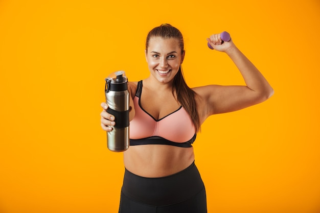 Portret van Europese mollige vrouw in sportieve beha bedrijf thermos met water terwijl het opheffen van halter, geïsoleerd op gele achtergrond