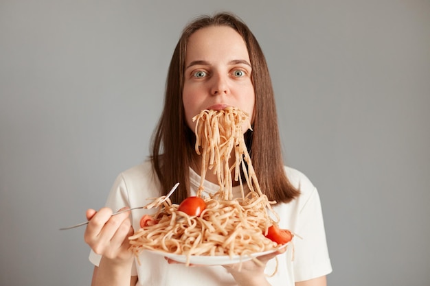 Portret van extreem hongerige vrouw met bruin haar die smakelijke pasta eet geïsoleerd over grijze achtergrond poserend met bord in handen kauwende noedels kijkend naar de camera