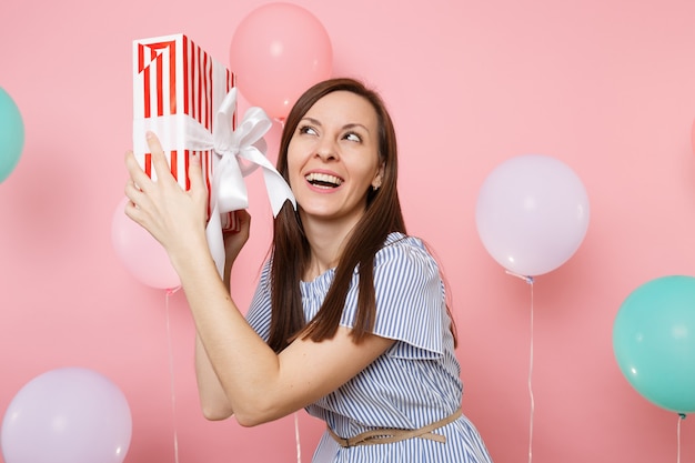 Portret van fascinerende lachende jonge vrouw in blauwe jurk met rode doos met cadeau aanwezig op pastelroze achtergrond met kleurrijke luchtballonnen. Verjaardagsfeestje, mensen oprecht emotie concept