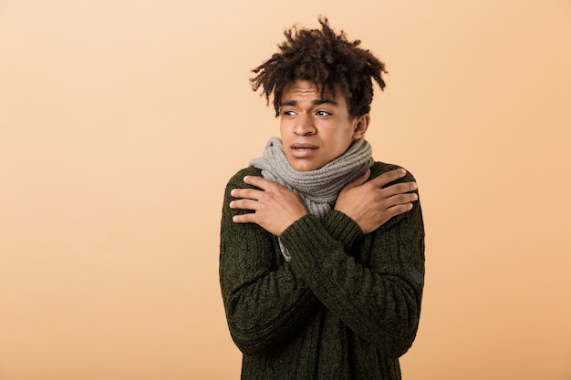 Portret van fronsende Afro-Amerikaanse man met trui en sjaal trillen als gevolg van koude, geïsoleerd over beige muur