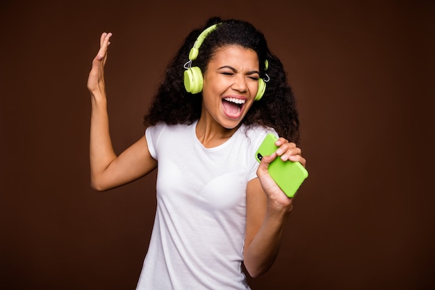 Portret van funky Afro-Amerikaanse jeugd stel je voor dat ze een rockconcert uitvoert, een liedje zingt, gebruik een smartphone-microfoon, luister naar muziek op een groene draadloze headset, draag een wit t-shirt.
