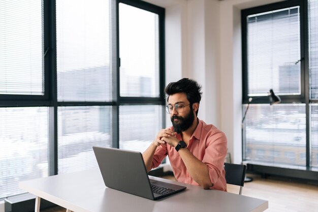 Portret van gefocuste Indiase zakenman met bril die online praat via videogesprek op laptop zittend aan bureau bij raam op zoek naar scherm Freelancer-man die computer gebruikt voor videogesprek