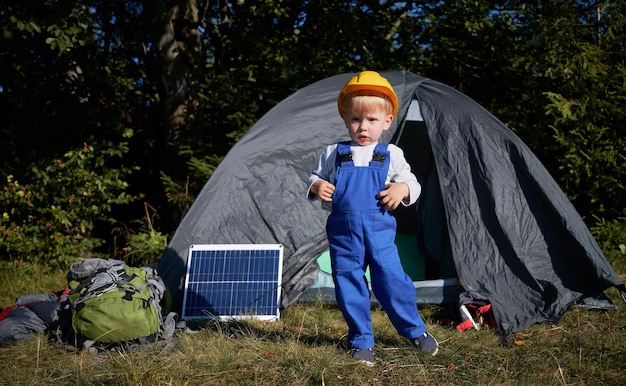 Portret van gefocuste kleine jongen in bouwhelm staande op gras voor tent één zonnepaneel en rugzak voor wandeling Jonge generatie die nieuwe zonne-energie demonstreert