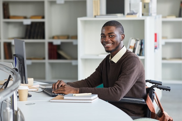 Portret van gehandicapte Afro-Amerikaanse man met behulp van computer en glimlachend in de camera tijdens het studeren in de universiteitsbibliotheek,