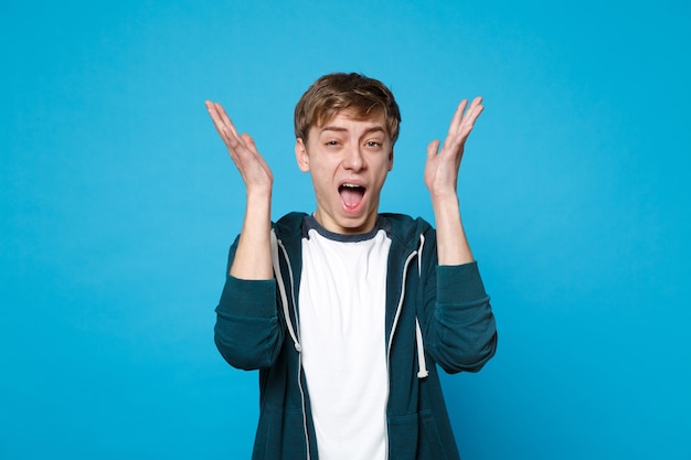 Portret van geïrriteerde jonge man in casual kleding schreeuwen, opstaan en handen verspreiden geïsoleerd op blauwe muur. mensen oprechte emoties, lifestyle concept.