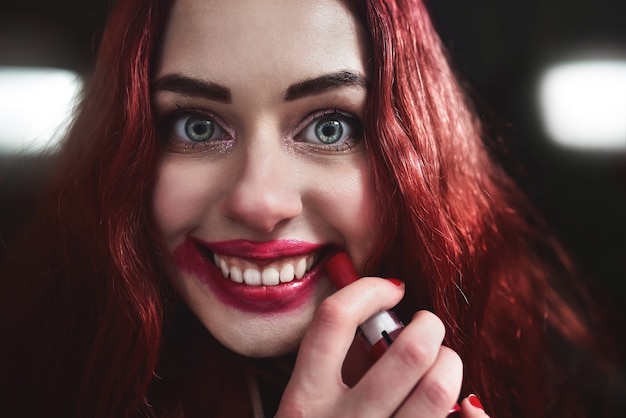 Portret van gek uitziende tiener meisje met rood haar, ze smeert rode lippenstift op haar gezicht, horror concept