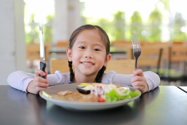 Portret van gelukkig Aziatisch kindmeisje die Varkensvleeslapje vlees en plantaardige salade op de lijst eten