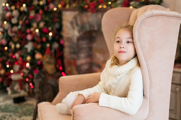 portret van gelukkig blond kind meisje in witte trui aanbrengen op de vloer in de buurt van de kerstboom