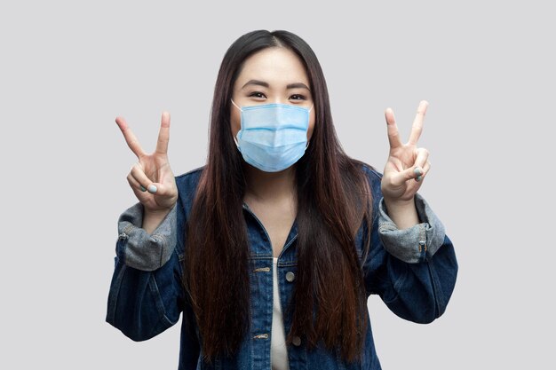Portret van gelukkig grappig mooi brunette Aziatisch meisje met medisch masker in blauw spijkerjasje dat staat en naar de camera kijkt met overwinnings- of vredesteken. studio opname, geïsoleerd op een grijze achtergrond.