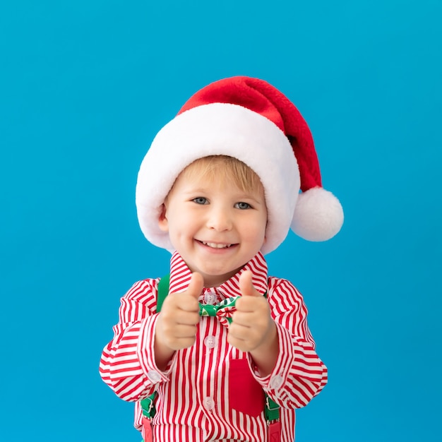 Portret van gelukkig kind duimen opdagen. Grappige jongen gekleed Kerstman hoed tegen blauwe achtergrond. Kerst vakantie concept