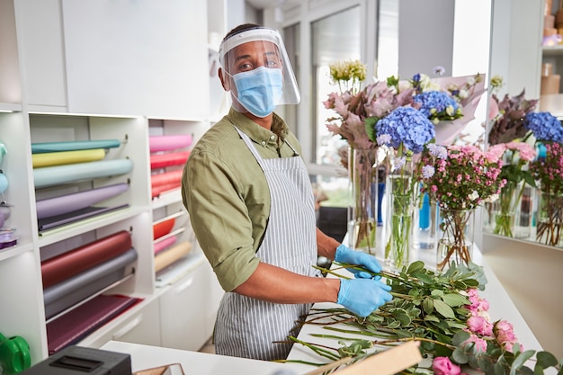 Portret van gelukkig knap mannetje in masker en glas makende rozenboeketten in winkel tijdens pandemie