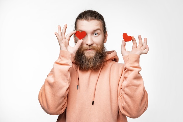 Portret van gelukkig knappe man met gember kapsel en baard op grijze achtergrond. Aantrekkelijke scandinavische man die speels is en rode harten voor zijn ogen houdt