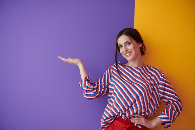 Portret van gelukkig lachende jonge mooie vrouw in een presenterend gebaar met open handpalm geïsoleerd op paarse en gele achtergrond. Vrouwelijk model in moderne modieuze kleding poseren in de studio